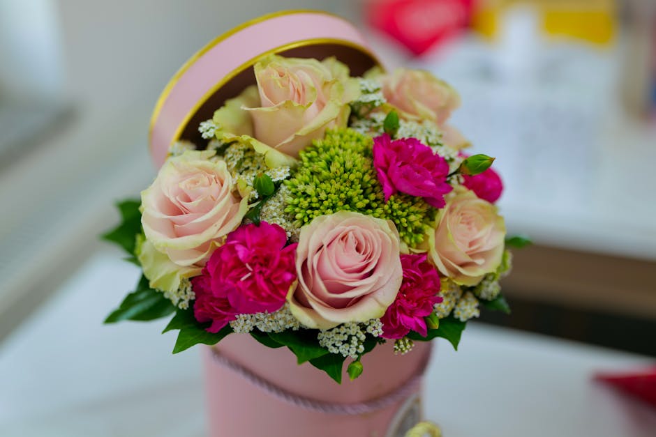 A round floral bouquet arrangement featuring pale pink roses, bright pink carnations, clusters of small white and green filler flowers, and lush green leaves, presented in a pastel pink cylindrical bo