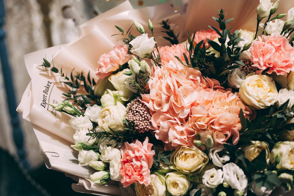 A beautifully arranged bouquet of fresh flowers featuring soft pink hydrangeas, white roses, and pale peach carnations, complemented by dark green foliage and small pink fillers. The bouquet is wrappe