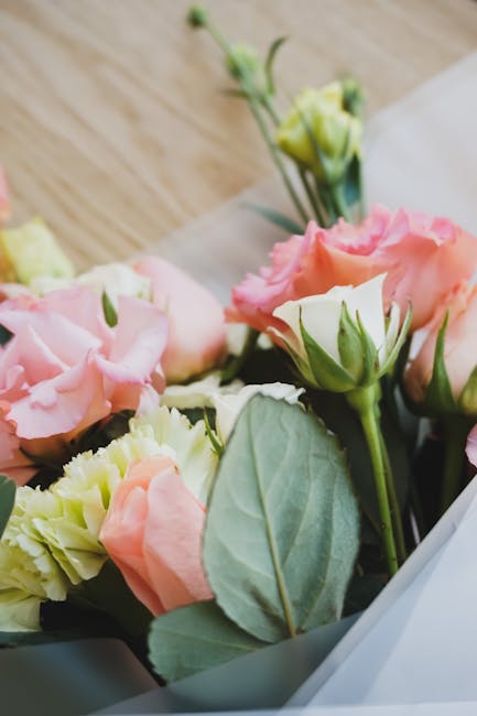A close-up of a bouquet featuring soft pink and pale yellow roses with multiple layers of petals, complemented by light green leaves and buds, arranged in a fresh floral bouquet. The flowers are wrapp