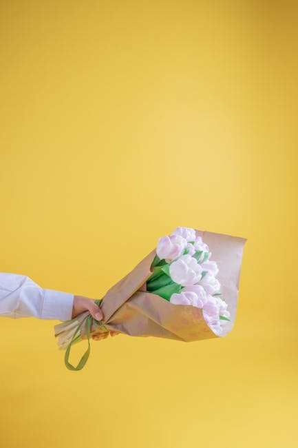 A person wearing a white sleeve holds a bouquet of fresh, pale pink tulips wrapped in light brown kraft paper with green ribbon against a vibrant yellow background. The tulips have open petals with a