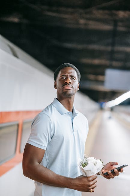 A young man with dark skin and short curly hair standing on a platform, holding a bouquet of white flowers wrapped in paper in his left hand and a smartphone in his right hand. He is wearing a light b