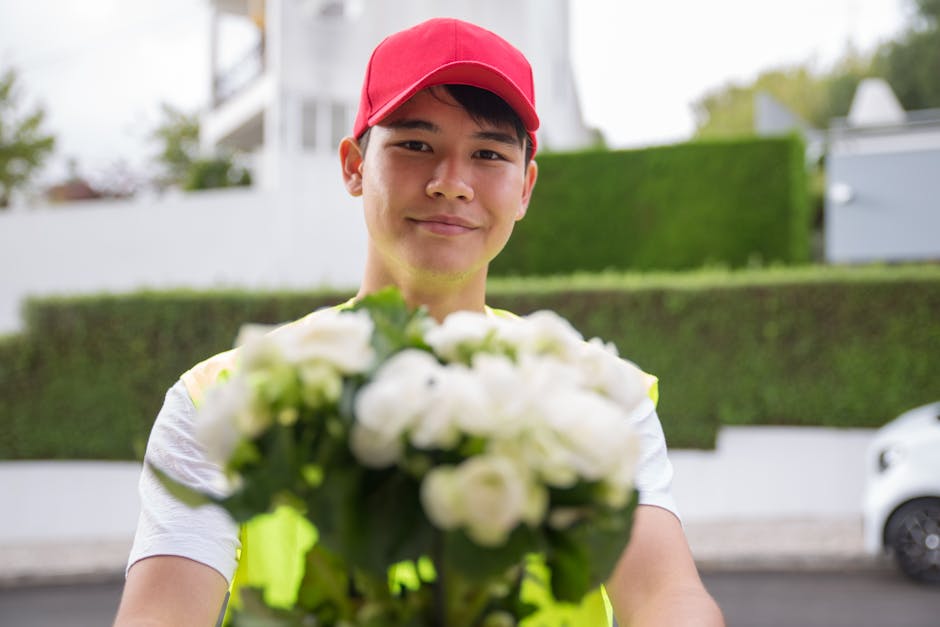 A delivery person wearing a red cap and a high-visibility vest is holding a bouquet of fresh white flowers, including roses and other blooms, wrapped in clear cellophane. The floral arrangement appear