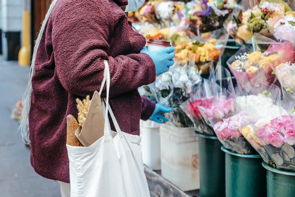 A person wearing a maroon fleece jacket and blue gloves is holding a plastic cup, standing in front of a floral display of fresh flower bouquets. The bouquets include pink roses, white blooms, and yel