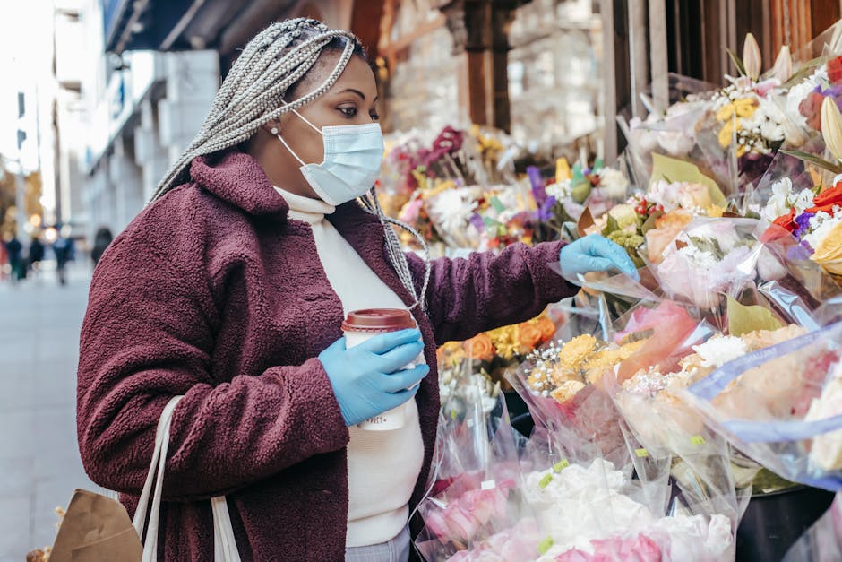 A woman wearing a light blue face mask, light blue gloves, and a white turtleneck sweater under a mauve fuzzy jacket selects a floral arrangement from an outdoor flower display along Main Road Gidea P